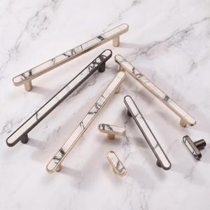 May include: A collection of cabinet hardware including long drawer pulls and small knobs. Each piece features a white marble inlay with grey veining, set into either a brushed gold or dark bronze metal frame with a smooth, elongated oval shape.
