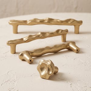 May include: A collection of gold-toned cabinet hardware featuring three elongated drawer pulls and one circular knob. Each piece has an irregular, textured surface with organic, undulating contours and a matte finish, showing mounting holes.