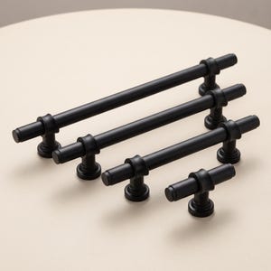 May include: A set of four matte black cabinet pulls of varying lengths, each featuring a cylindrical bar design with two circular mounting posts and flat end caps. The hardware is arranged in a diagonal line from the longest to the shortest piece.