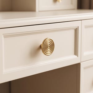 May include: A circular gold-toned metal cabinet knob featuring a concentric ring pattern on its face. The knob is mounted on a smooth, off-white drawer front with a rectangular recessed panel design, part of a larger piece of furniture.