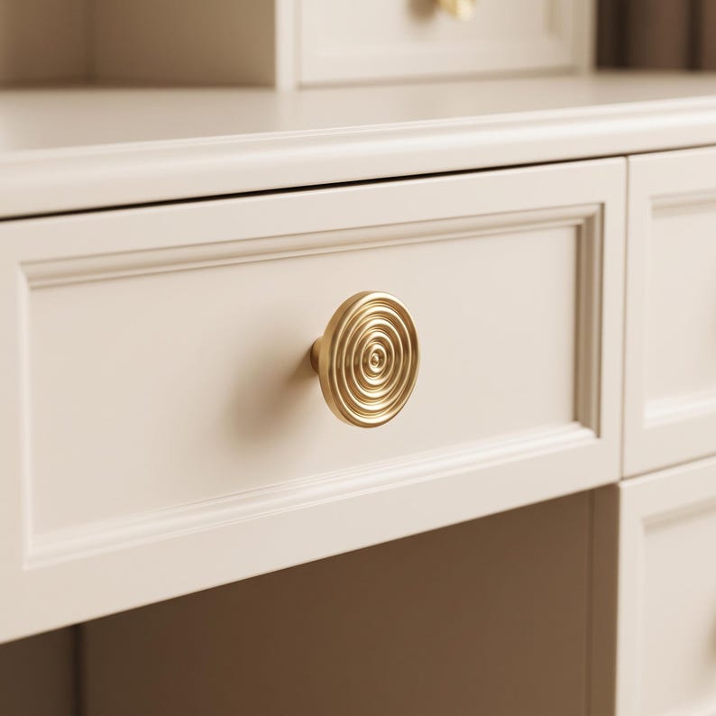 May include: A circular gold-toned metal cabinet knob featuring a concentric ring pattern on its face. The knob is mounted on a smooth, off-white drawer front with a rectangular recessed panel design, part of a larger piece of furniture.