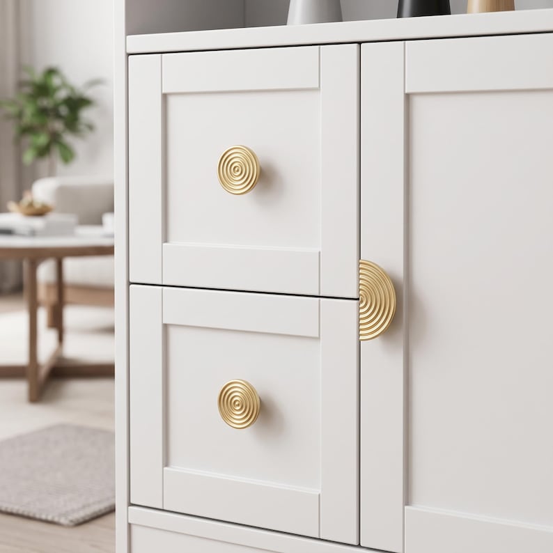 May include: A white cabinet featuring two stacked square drawers and a side door. The drawers are accented with circular gold knobs, while the door features a semi-circular gold handle. Each handle and knob displays a concentric ring pattern.