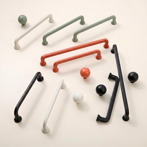 May include: A collection of cabinet hardware including various drawer pulls and round knobs in matte finishes. The set features colors such as white, sage green, terracotta orange, and black, showcasing smooth, rounded designs for furniture fixtures.