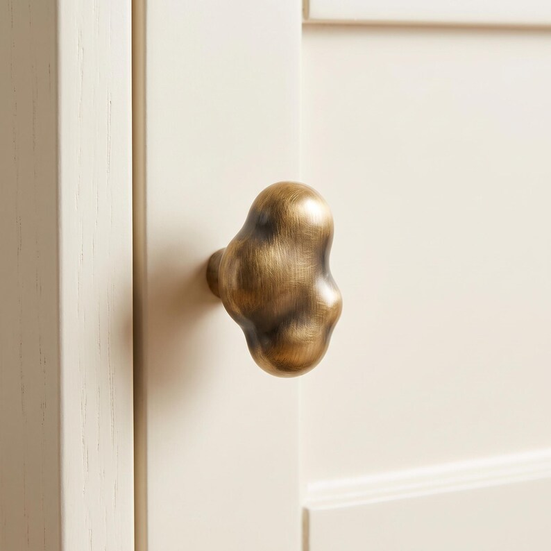 Op de afbeelding: Een messing kastknop met een onregelmatige, organische, wolkachtige vorm en een geborstelde metalen afwerking. De knop is gemonteerd op een licht cr&egrave;mekleurige houten kastdeur met een zichtbaar verticaal houtnerfpatroon op de rand.