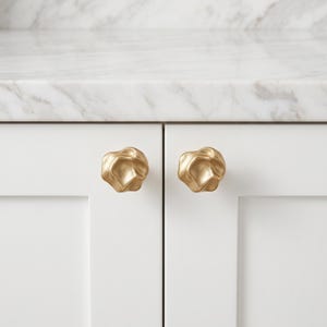 May include: Two gold-colored cabinet knobs with an irregular, organic, and textured surface. Each knob features a rounded, multi-faceted shape with deep indentations and raised edges, mounted onto the front of two white cabinet doors.