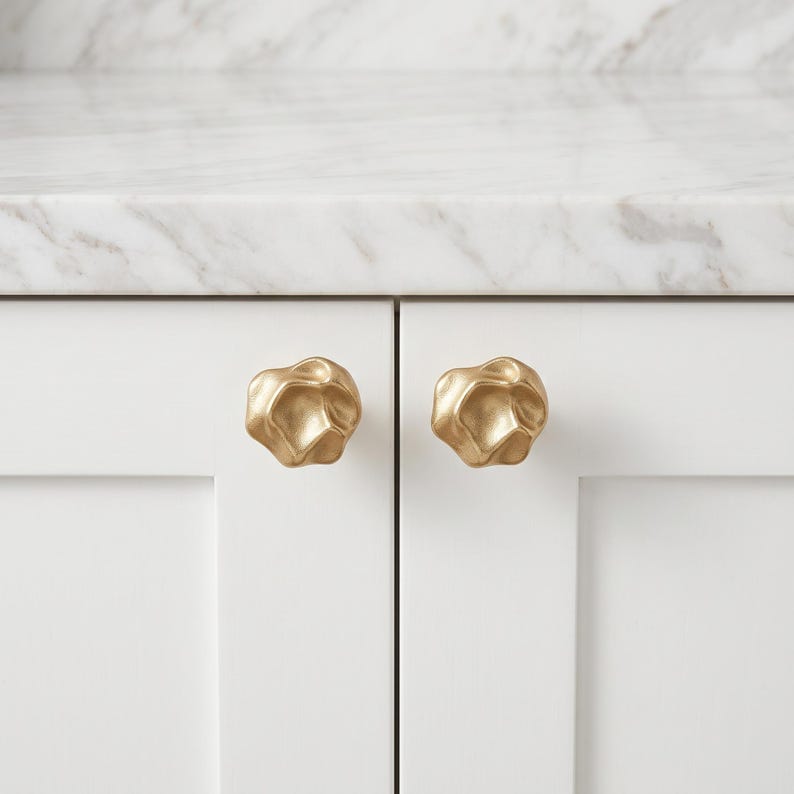 May include: Two gold-colored cabinet knobs with an irregular, organic, and textured surface. Each knob features a rounded, multi-faceted shape with deep indentations and raised edges, mounted onto the front of two white cabinet doors.