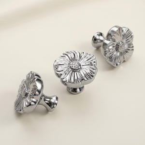 May include: Three silver-toned metal cabinet knobs shaped like flowers. Each knob features a central textured circular core surrounded by smooth, radiating petals. The metallic surface is reflective and polished, showing a high-gloss finish.