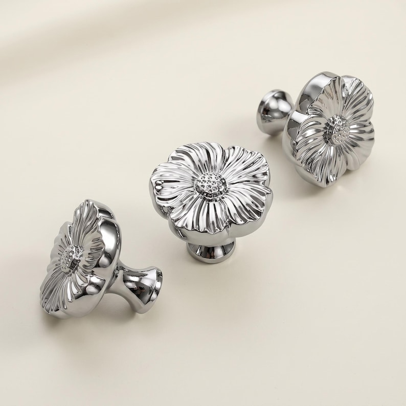 May include: Three silver-toned metal cabinet knobs shaped like flowers. Each knob features a central textured circular core surrounded by smooth, radiating petals. The metallic surface is reflective and polished, showing a high-gloss finish.