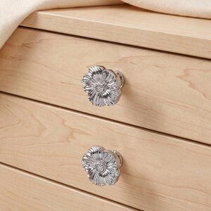 May include: Two silver-toned metal drawer knobs shaped like flowers with detailed petals and a textured centre. The knobs are attached to a light-coloured wooden drawer front with a visible grain pattern, featuring a smooth, polished metallic finish.