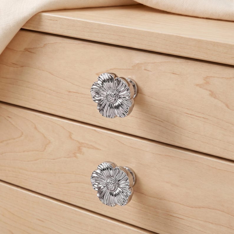 May include: Two silver-toned metal drawer knobs shaped like flowers with detailed petals and a textured centre. The knobs are attached to a light-coloured wooden drawer front with a visible grain pattern, featuring a smooth, polished metallic finish.