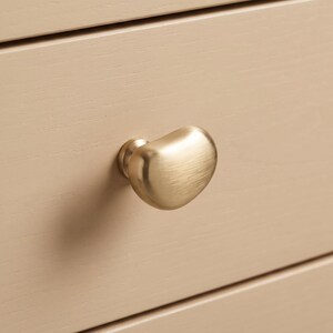 Op de afbeelding: Een geborstelde gouden meubelknop met een organische, kiezelachtige vorm bevestigd aan een lichtbeige houten ladefront. Het metalen beslag heeft een zachte, matte afwerking en een afgeronde, asymmetrische vorm die uitsteekt.