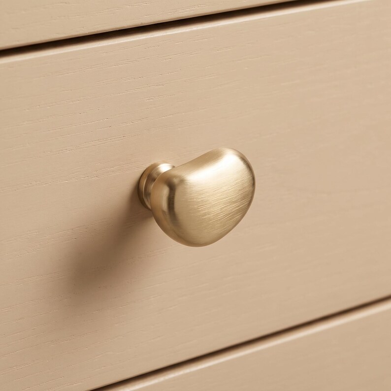 Op de afbeelding: Een geborstelde gouden meubelknop met een organische, kiezelachtige vorm bevestigd aan een lichtbeige houten ladefront. Het metalen beslag heeft een zachte, matte afwerking en een afgeronde, asymmetrische vorm die uitsteekt.