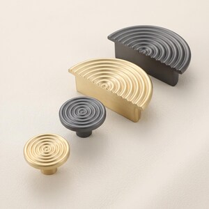 May include: A collection of four cabinet hardware pieces featuring two circular knobs and two semi-circular pulls. The items are finished in matte gold and matte black, each displaying a concentric ripple pattern across the top surface.
