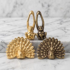 May include: Two brass monkey figurines with arms raised overhead and two brass peacock figurines with fanned tail feathers. One monkey and one peacock have a bright polished gold finish, while the other pair features a darker, antique brass patina.