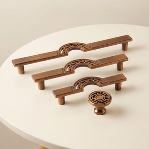 Op de afbeelding: Drie bronskleurige metalen ladehandgrepen van verschillende lengtes en een bijpassende ronde knop. Elk stuk heeft een centrale halfronde uitsparing met een ingewikkeld geometrisch patroon. De hardware heeft een gladde, geborstelde afwerking.
