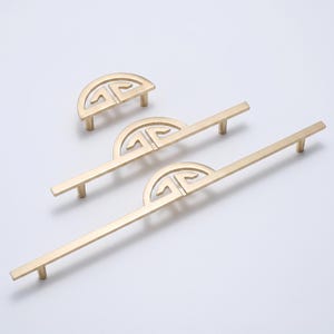 May include: Three gold-toned metal cabinet pulls of varying lengths, each featuring a central semi-circular decorative element with a cut-out geometric pattern and two cylindrical mounting posts extending from the base of the handle.
