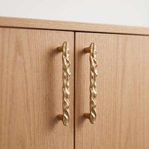 May include: Two vertical cabinet handles made of brushed gold-toned metal with an irregular, wavy, and textured surface. These handles are mounted onto a light-colored wooden cabinet door with a visible grain pattern, featuring a smooth finish.