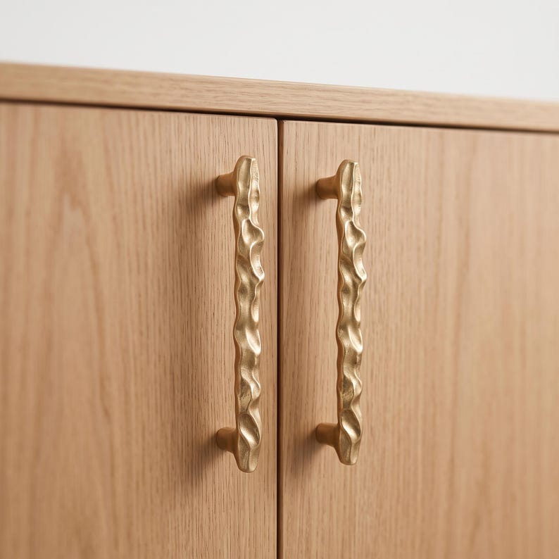 May include: Two vertical cabinet handles made of brushed gold-toned metal with an irregular, wavy, and textured surface. These handles are mounted onto a light-colored wooden cabinet door with a visible grain pattern, featuring a smooth finish.