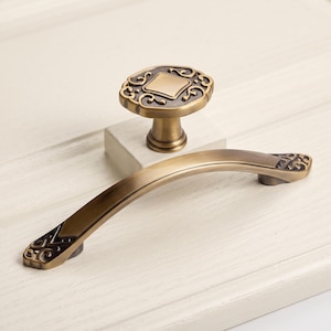 Pode incluir: Um puxador de móvel em metal tom latão com centro quadrado e volutas ornamentais, emparelhado com um puxador de gaveta curvo a condizer, apresentando detalhes decorativos escuros em ambas as extremidades e um acabamento metálico escovado.