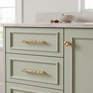 May include: A sage green cabinet featuring two horizontal drawers and one door. The drawers are fitted with long, textured gold-toned metal pulls, while the cabinet door features a single, small, rounded, textured gold-toned metal knob.