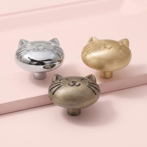 Puede incluir: Tres pomos de metal para armarios con forma de cabeza de gato, con caras sonrientes y bigotes. Un pomo tiene un acabado plateado pulido, otro un acabado dorado cepillado y el tercero un acabado de bronce antiguo oscuro.
