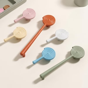 May include: A collection of cabinet hardware featuring a spiral lollipop design in various colors including pink, orange, beige, white, blue, and sage green. The set includes both short-handled knobs and longer, elongated drawer pulls.