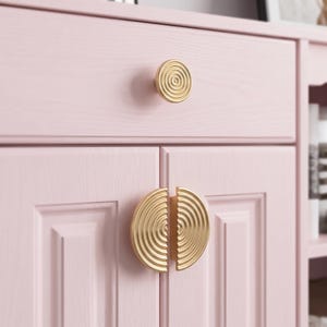 May include: A light pink wooden cabinet featuring a single circular gold knob on the top drawer and two semi-circular gold handles on the lower cabinet doors. Each gold handle displays a concentric ring pattern etched into the metal surface.
