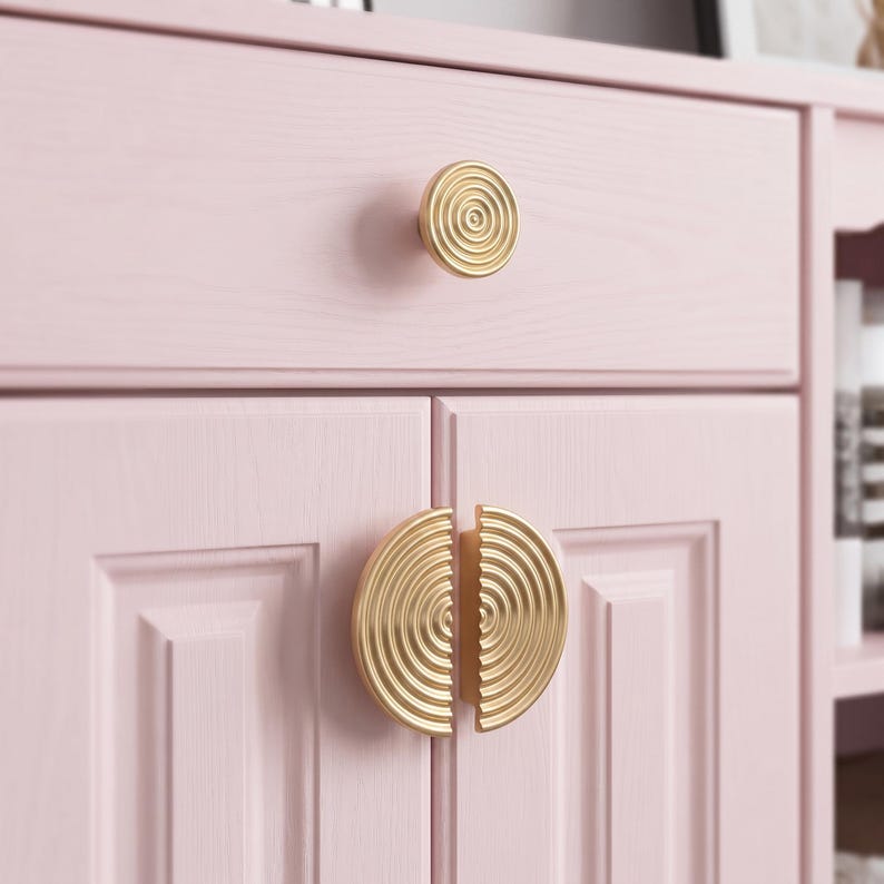 May include: A light pink wooden cabinet featuring a single circular gold knob on the top drawer and two semi-circular gold handles on the lower cabinet doors. Each gold handle displays a concentric ring pattern etched into the metal surface.