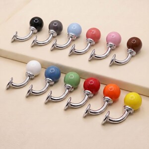 May include: A collection of twelve wall hooks, each featuring a smooth, spherical knob in a different solid color, including black, gray, light blue, pink, white, dark blue, green, red, orange, yellow, and brown, attached to a curved chrome arm.