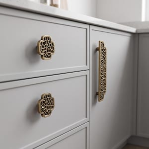 May include: Two square-shaped brass cabinet knobs with rounded corners and a geometric honeycomb-like cutout pattern, alongside a matching long rectangular brass cabinet pull handle, all mounted on light grey kitchen cabinetry drawers.
