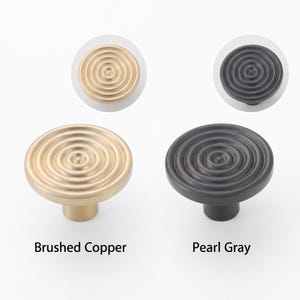 May include: Two round cabinet knobs with a concentric circle pattern. One knob is finished in brushed copper and the other in pearl gray. Each knob features a central point with radiating rings. The text Brushed Copper and Pearl Gray is visible.