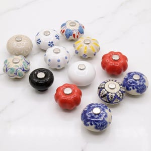 Op de afbeelding: Een collectie van dertien ronde keramische kastknoppen met diverse ontwerpen, waaronder effen kleuren zoals rood, zwart en wit, naast bloemen-, stippen- en bladpatronen, elk voorzien van een kleine zilverkleurige metalen schroef in het midden.