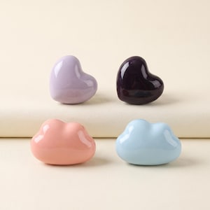 May include: Four ceramic drawer knobs in a smooth, glossy finish. Two knobs are heart-shaped in light purple and dark purple, while the other two are cloud-shaped in soft peach and light blue. These decorative hardware pieces have a rounded profile.