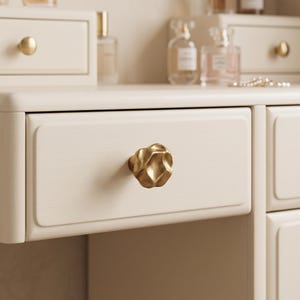 May include: A gold-toned cabinet knob with an irregular, organic, and textured surface. The knob is mounted on a light cream-colored wooden drawer front. The finish of the knob is matte and metallic, contrasting with the smooth, light-colored wood.