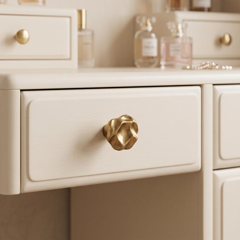 May include: A gold-toned cabinet knob with an irregular, organic, and textured surface. The knob is mounted on a light cream-colored wooden drawer front. The finish of the knob is matte and metallic, contrasting with the smooth, light-colored wood.