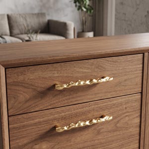 May include: A wooden dresser featuring two drawers with horizontal grain patterns. Each drawer is equipped with a long, gold-colored metal handle that has an irregular, wavy, and textured surface design across its entire length.