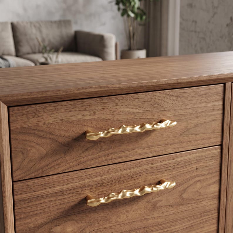 May include: A wooden dresser featuring two drawers with horizontal grain patterns. Each drawer is equipped with a long, gold-colored metal handle that has an irregular, wavy, and textured surface design across its entire length.