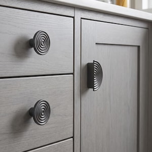 May include: Two circular cabinet knobs and one semi-circular cabinet pull, all featuring a matte black finish with a concentric ring pattern. These hardware pieces are mounted on light grey wooden drawers and a cabinet door with a visible grain.