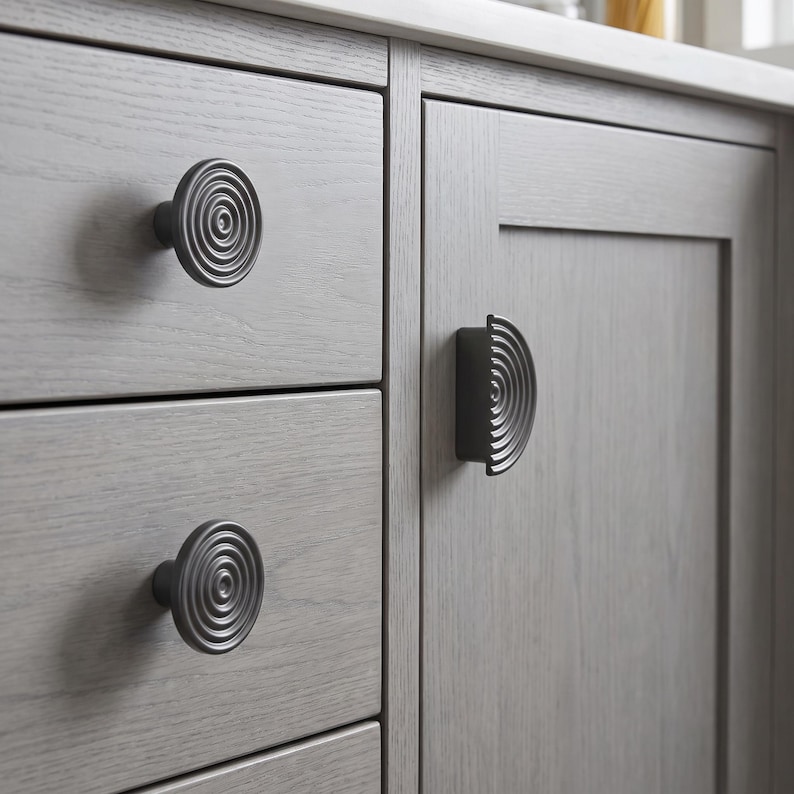 May include: Two circular cabinet knobs and one semi-circular cabinet pull, all featuring a matte black finish with a concentric ring pattern. These hardware pieces are mounted on light grey wooden drawers and a cabinet door with a visible grain.