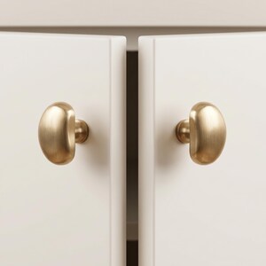 Op de afbeelding: Twee kastknoppen van geborsteld messing met een gladde, afgeronde, half-ovale vorm. Elke knop is gemonteerd op een ronde basis en bevestigd aan een plat, gebroken wit kastdeurpaneel. De metalen afwerking heeft een zachte, matte, goudkleurige textuur.