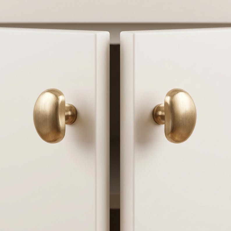 Op de afbeelding: Twee kastknoppen van geborsteld messing met een gladde, afgeronde, half-ovale vorm. Elke knop is gemonteerd op een ronde basis en bevestigd aan een plat, gebroken wit kastdeurpaneel. De metalen afwerking heeft een zachte, matte, goudkleurige textuur.