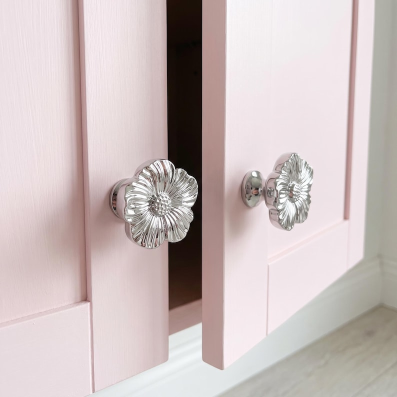 May include: Two silver-toned metal cabinet knobs shaped like flowers with textured petals and a central stamen detail, mounted on a pair of light pink cabinet doors with a flat panel design and a visible vertical seam between the two doors.