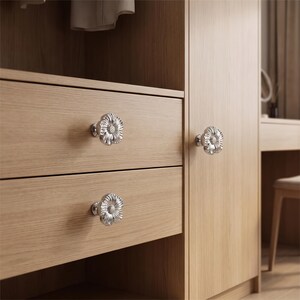 May include: Three silver-toned metal cabinet knobs shaped like flowers with detailed petals and a textured centre. These knobs are mounted on light-coloured wooden drawers and a cabinet door, featuring a polished finish that reflects light.