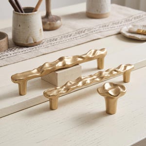 May include: Two gold-toned metal drawer pulls with an irregular, wavy, organic texture and a single matching gold-toned metal cabinet knob with a rounded, undulating top surface, all featuring a matte finish and cylindrical mounting posts.