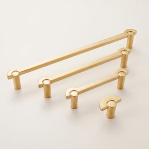 May include: A set of four gold-toned metal hardware pieces consisting of three linear drawer pulls of varying lengths and one single semi-circular knob. Each pull features a straight bar with cylindrical mounting posts and curved ends.