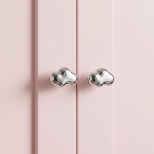 Op de afbeelding: Twee zilverkleurige kastknoppen in de vorm van gestileerde wolken, elk met een glad, reflecterend metalen oppervlak. Deze wolk-vormige hardware is gemonteerd op een vlakke, lichtroze kastdeur met verticale paneeldetails.