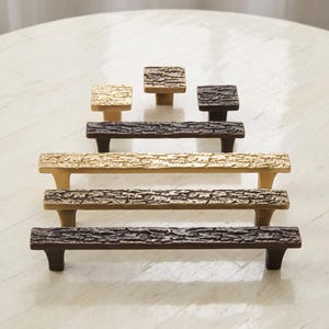 May include: A collection of cabinet hardware featuring three square knobs and three rectangular pulls. The pieces have a textured, bark-like surface finish and are available in metallic gold and dark bronze tones, arranged in a tiered formation.