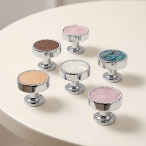 May include: Six circular cabinet knobs with silver-toned metal bases and beaded edges. The tops feature varied inlays including light wood, dark wood, iridescent abalone shell, white mother-of-pearl, and two pieces of smooth, pale pink stone.