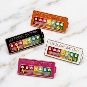 Puede incluir: Cuatro pines de esmalte rectangulares con forma de batería, cada uno con el texto MY SOCIAL BATTERY y un indicador deslizante con un icono de rayo. Los pines son de color naranja, negro, blanco y rosa, con caras de humor coloridas.