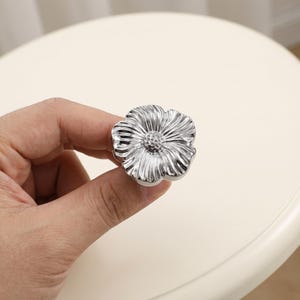 May include: A shiny silver-toned cabinet knob shaped like a flower with five distinct petals and a textured, dotted centre. The metallic surface reflects light, highlighting the radial grooves on each petal and the rounded, organic floral form.